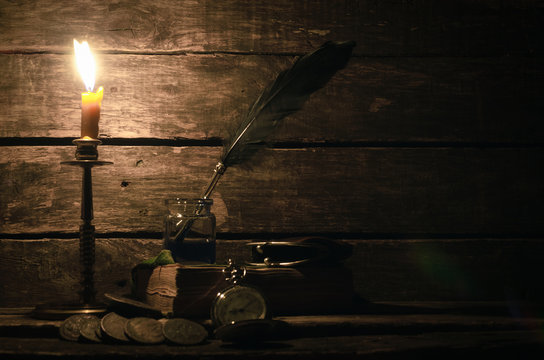 Quill Pen In A Inkpot, Wallet, Pocket Watch, Ancient Coins And Book In The Light Of Burning Candle On The Writer Desk Background. Education Concept.