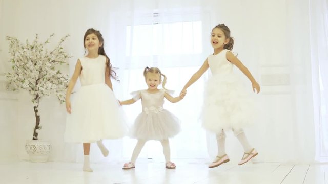 Three beautiful little girls are holding hands and dancing on the stage. Children are dressed in lush long dresses, they look like little brides or ballerinas. Girls hold hands and bounce up. Stylish