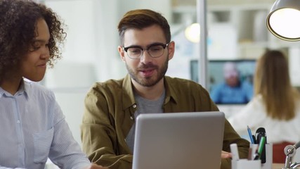 Medium shot of two young multi ethnic office workers sitting at desk and using laptop computer together - Powered by Adobe