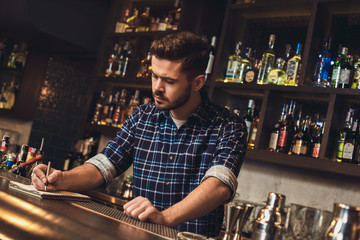 Young bartender standing at bar counter writing receipt concentrated