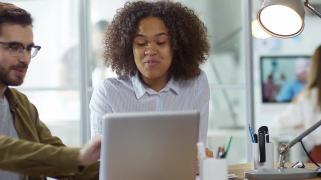 Tracking shot of two optimistic young colleagues of different ethnicities sitting at office desk and discussing something using laptop computer