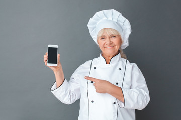 Senior woman chef studio standing isolated on gray with smartphone showing app on screen looking camera joyful