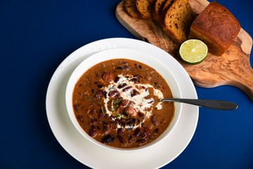 Bean soup with sliced sausages, dried plums and cream in white plate,bread on board, all on blue background.