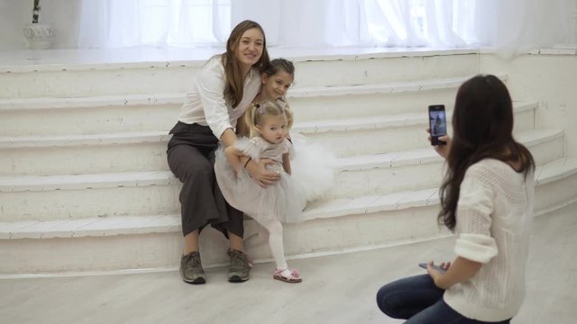 Mom is trying to take pictures with smart daughters. The girls sit in her arms, a friend photographs on the phone. But the little displeased babe runs away from the frame.