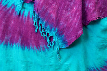 Close up of multi colored tie dyed fabric background cloth hanging showing corner edge and tassels, pink and purple on aqua blue.