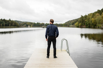 A pensive bearded man wearing a blue jacket and jeans, standing outside with a big lake with surrounding woods.