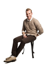 Handsome blonde man wearing a brown pullover and white shirt, sitting down laughing against a white background.