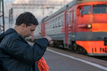 A man wipes his glasses with a cloth, sitting on the platform on the background of standing next to...