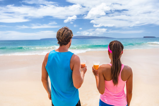 Healthy people active lifestyle. Fitness runners couple on beach drinking breakfast fruit juice in smoothie cups looking at ocean. Summer background.