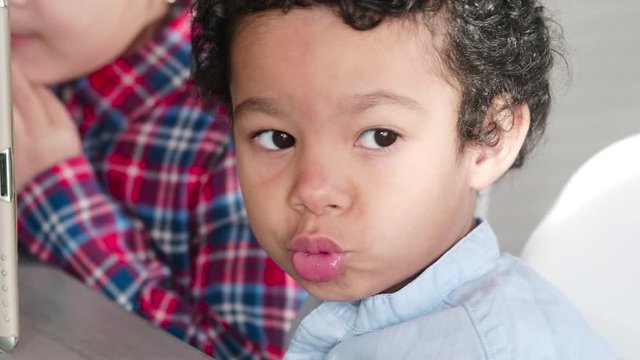 Close-up of cute little African American boy looking at side and pouting his lips while girl sitting next to him and using tablet