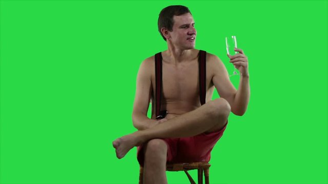Young sexy man with a beautiful muscular body is resting sitting on a deck chair against a green screen. Man resting with a glass of wine on green background.
