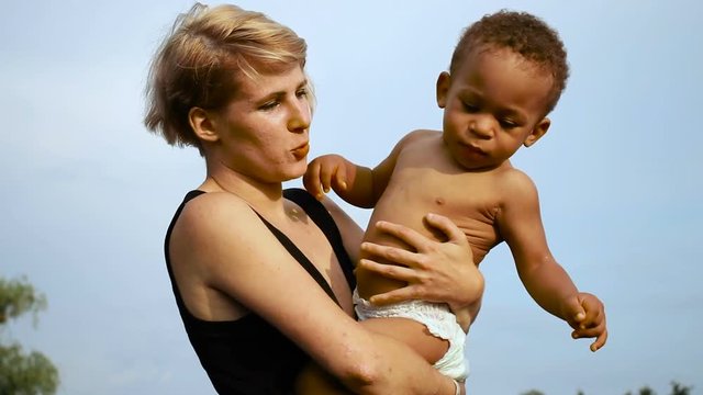 Happy multiracial family. Young mother holds her little son in her arms, hugs him and kisses him, in the summer park against the background of blue sky. 