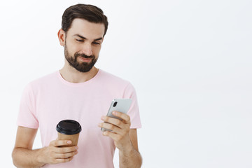 Carefree happy and calm attractive european man with beard and cool hairstyle holding smartphone and paper cup of coffee smirking with delight while gazing at device screen over gray wall