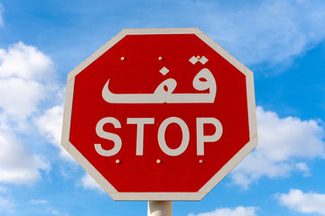 Stop sign in English and Arabic with a blue sky and cloudy background.