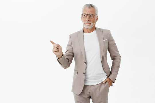 Unretouched Shot Of Good-looking Intelligent And Elegant Old Man With Grey Beard And Hair Holding Hand In Pocket Pointing And Looking At Upper Left Corner With Questioned Uncertain And Disliking Look