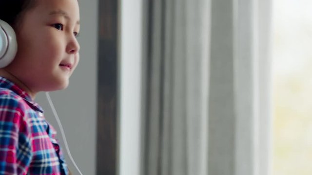 Tilt up shot of playful little Asian girl in headphones listening to music on smartphone and dancing at home