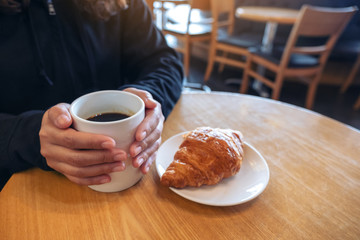 A woman holding coffee cup with a piece of croissant in a white plate on wooden table in cafe