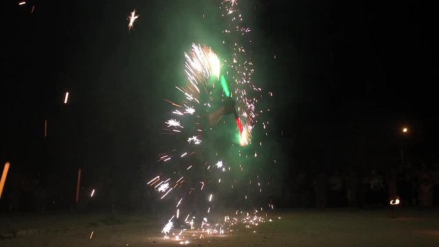 Three fire show performers standing in fire circle, performing. One juggles burning sticks, other flails them - slow motion