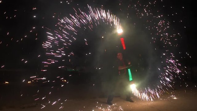 Three fire show performers standing in fire circle, performing. One juggles burning sticks, other flails them - slow motion