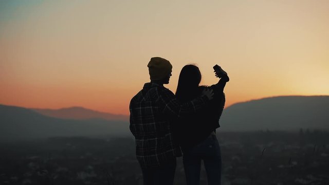 Silhouette of young couple in love enjoying a sunset over the mountains. They taking photo on smartphone. Vacation, travel concept