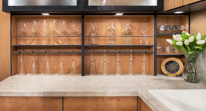 A bouquet of white tulips stands on a marble tabletop in the interior of a modern kitchen. Glass wine glasses and glasses are placed on wooden shelves.