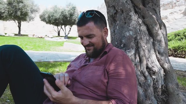 Happy young hipster man with smartphone, sitting on grass near tree in summer time.