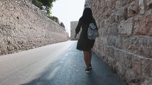 Young brunette with long hair walks down the street of Jerusalem and spending her hand over the stone wall of the old city.