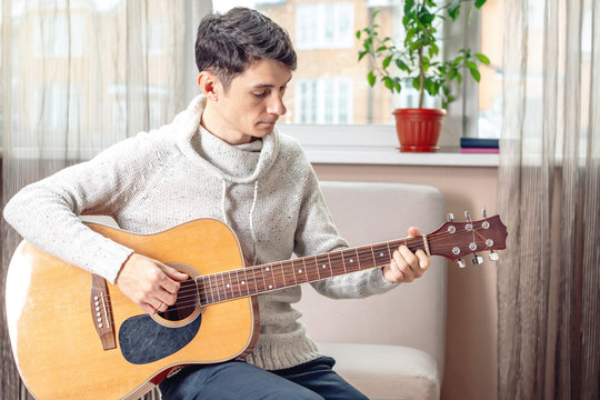 Young attractive male musician sitting on a chair playing acoustic guitar in room. Concept of music as a hobby