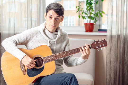 Young attractive male musician sitting on a chair playing acoustic guitar in room. Concept of music as a hobby