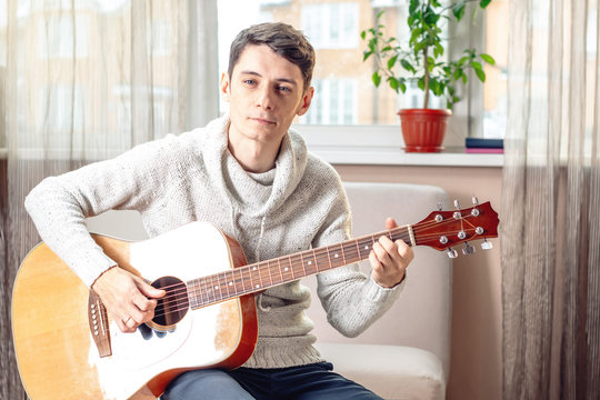 Young attractive male musician sitting on a chair playing acoustic guitar. Concept of music as a hobby