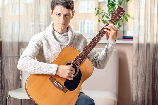 Young attractive male musician sitting on a chair playing acoustic guitar. Concept of music as a hobby