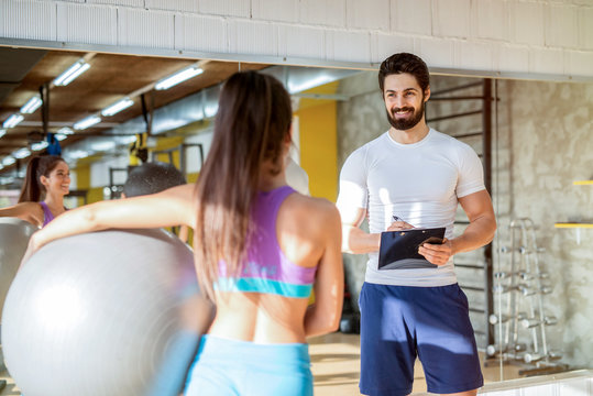 Picture of personal fitness trainer talking with his female client while she is holding big ball in her hands.