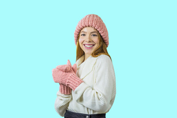 A young red-haired teenager girl in a knitted pink hat and mittens is standing in a half-turn with her arms folded smiling and looking away, the girl's emotions, winter holidays and holidays