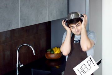 Funny loser man male guy with metal sieve, frying pan, wooden kitchen utensils and apron trying to cook, failed and need help on grey modern loft kitchen. Unsuccessful bachelor on kitchen concept.