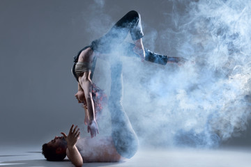 Obraz premium Couple in love dancers making dance element in smoke fume on isolated grey background scene. Redhead woman female dancer athlete gymnast in overalls with muscle male man in jeans and bare naked torso