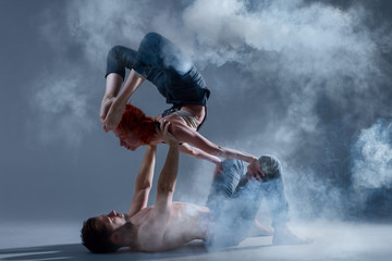 Couple in love dancers making dance element in smoke fume on isolated grey background scene. Redhead woman female dancer athlete gymnast in overalls with muscle male man in jeans and bare naked torso