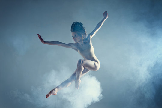 Dancing in cloud concept. Muscle brunette beauty female girl adult woman dancer athlete in dust / fog. Girl wearing dance bodysuit jumping in mid air, performance on isolated grey / black background