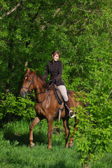 Beautiful equestrian women rides saddle horse in woods glade at sunset