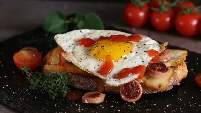 Pouring ketchup over open sandwich made with egg, cheese and sausages on slate
