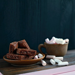 pink marshmallows and milk chocolate on a brown plate and a cup of coffee with marshmallows on a wooden background