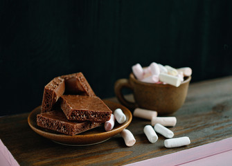 pink marshmallows and milk chocolate on a brown plate and a cup of coffee with marshmallows on a wooden background