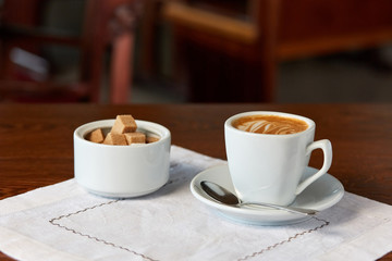 mug of latte with a foam pattern stands on a table on a white napkin, next to a sugar bowl with cane and beet sugar