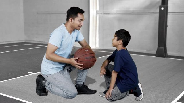 JAKARTA, Indonesia - January 08, 2019: Young father and his son talking together while holding a basketball and sitting in basketball court. Shot in 4k resolution