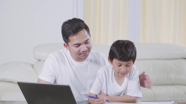 Young father helping his son to study with laptop and book on the table in the living room at home. Shot in 4k resolution
