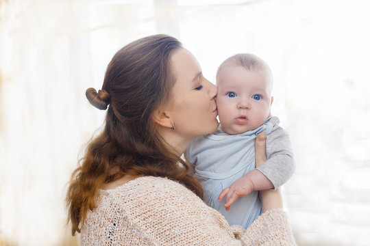 Gentle photo of mother and baby. Mom kisses her son on the cheeks on a light background