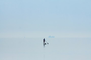 silhouette figure of a woman standing on a paddleboard with oar in her hands, still water surface, transport ship on horizon