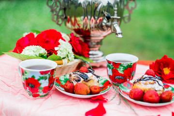Samovar with a cup of morning tea, cinnamon buns and strawberry on the table on nature background