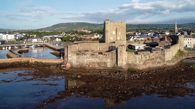 Medieval Norman Castle in Carrickfergus near Belfast in sunrise light. Aerial 4K flyby video with water reflection, marina, groin, breakwater, town and far view of Belfast in the background