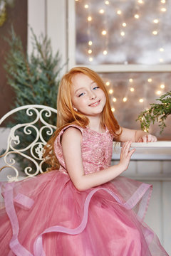 Girl with long red hair in a pink dress sitting at the table. Carnival holiday birthday. Portrait of a cheerful red-haired girl with big blue eyes