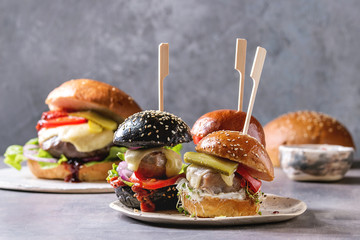 Variety of homemade classic and mini burgers in wheat and black buns with beef and veal cutlets, melted cheese and vegetables on white ceramic board over grey blue table. - obrazy, fototapety, plakaty